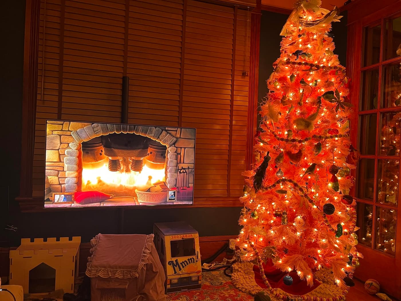 A cozy living room scene features a television displaying a crackling fireplace animation. Next to it stands a brightly lit orange Christmas tree adorned with colorful ornaments and decorations. The atmosphere is warm and festive, enhanced by soft lighting.