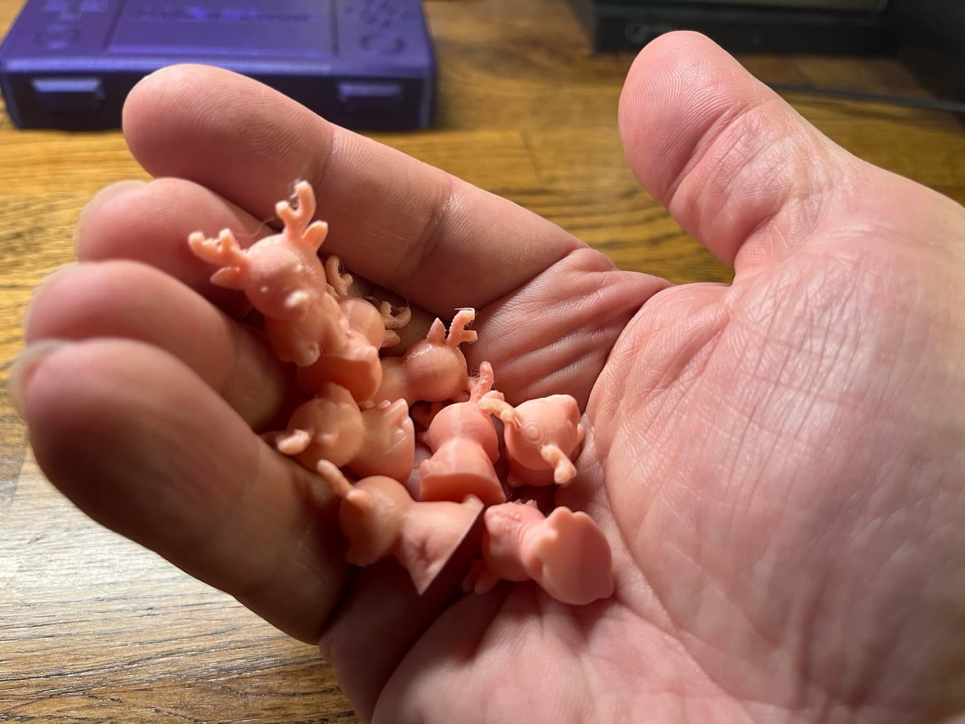 A hand holding several small, pink plastic figurines resembling cartoon reindeer, against a wooden surface with a purple object in the background.