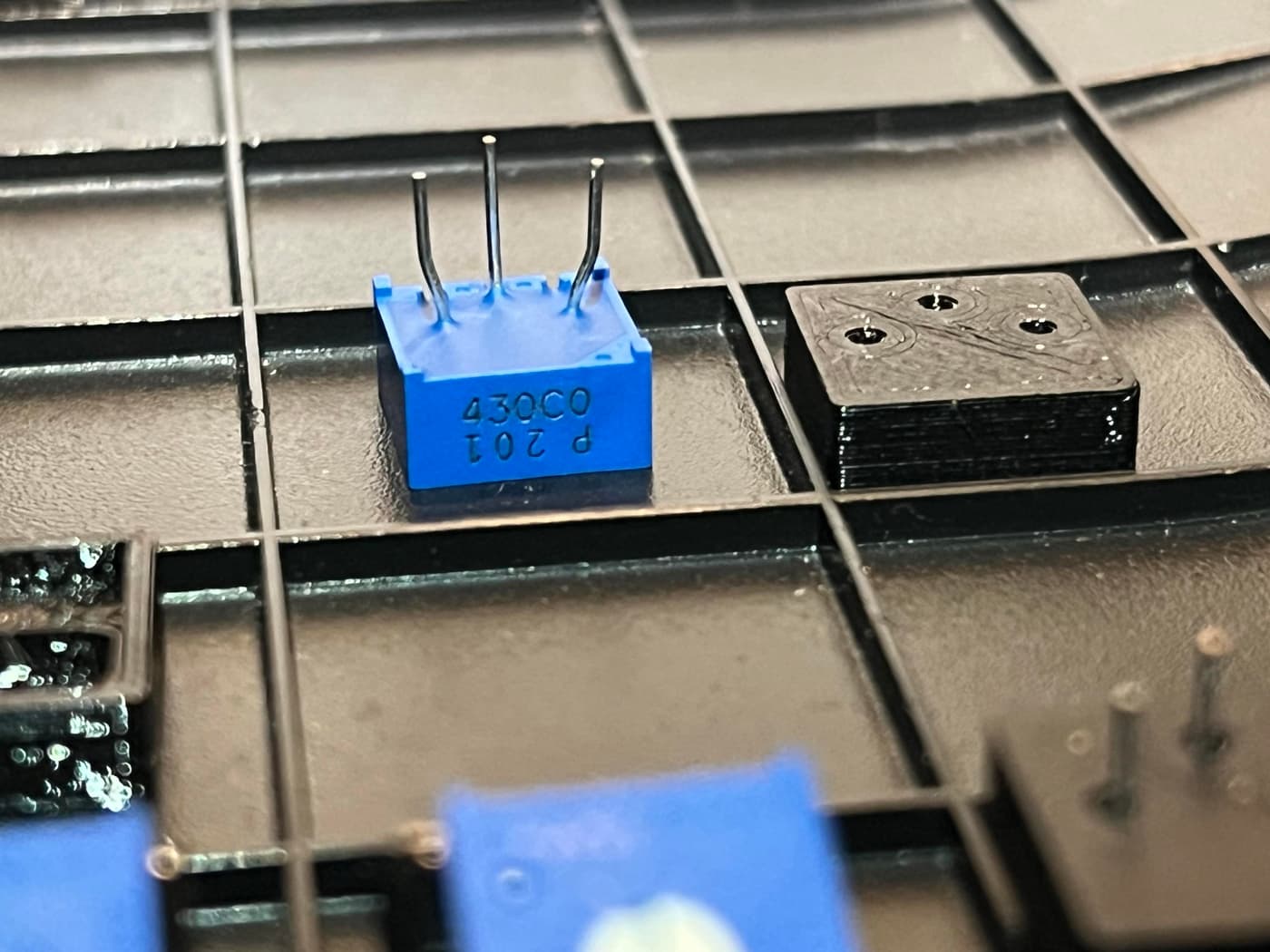 A blue electronic component with three bent metal pins on a black grid-like surface