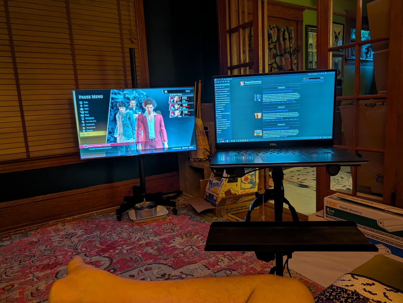 A bare leg in foreground, with a video game 'Pause Menu' on one screen and a blog on a laptop on another stand.