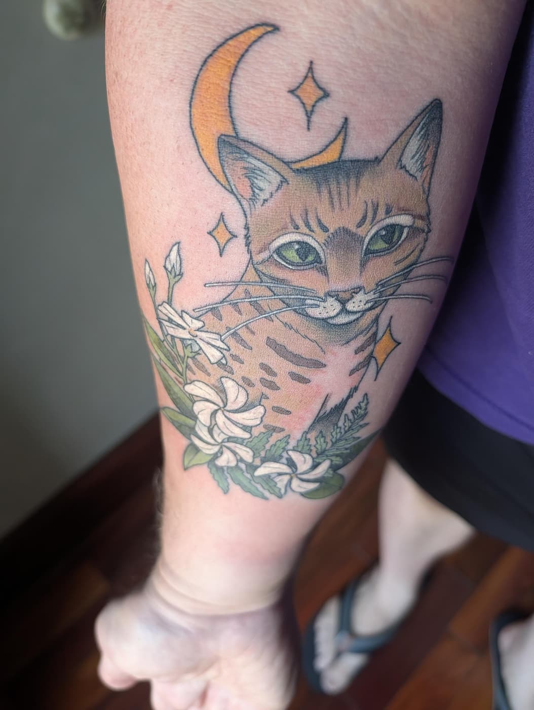 A colorful tattoo on an arm featuring a cat with green eyes, a crescent moon, stars, and white flowers.