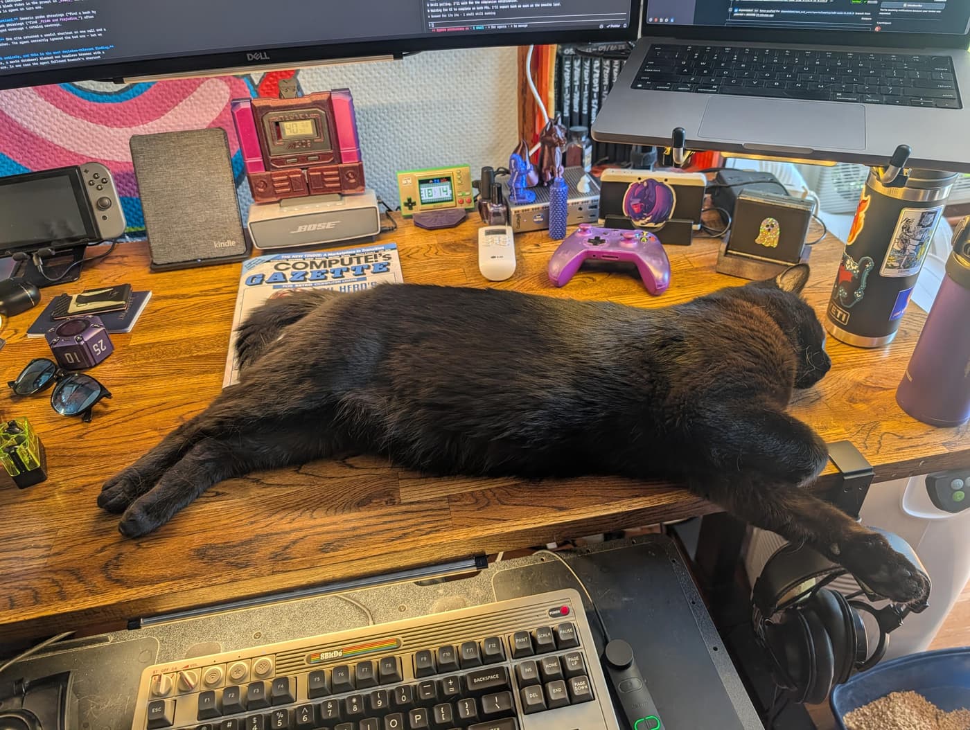 A black cat sleeps stretched out across a cluttered desk filled with computer monitors, a retro keyboard, and gaming accessories.